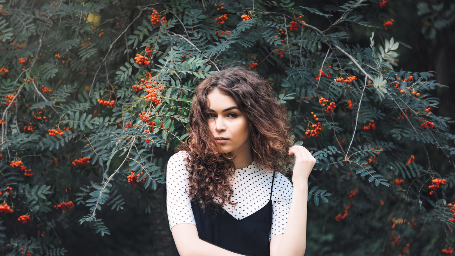 fashionable woman with curly brown hair wearing fashionable top and dress in front of mistletoe tree while gazing into the camera and teasing hair