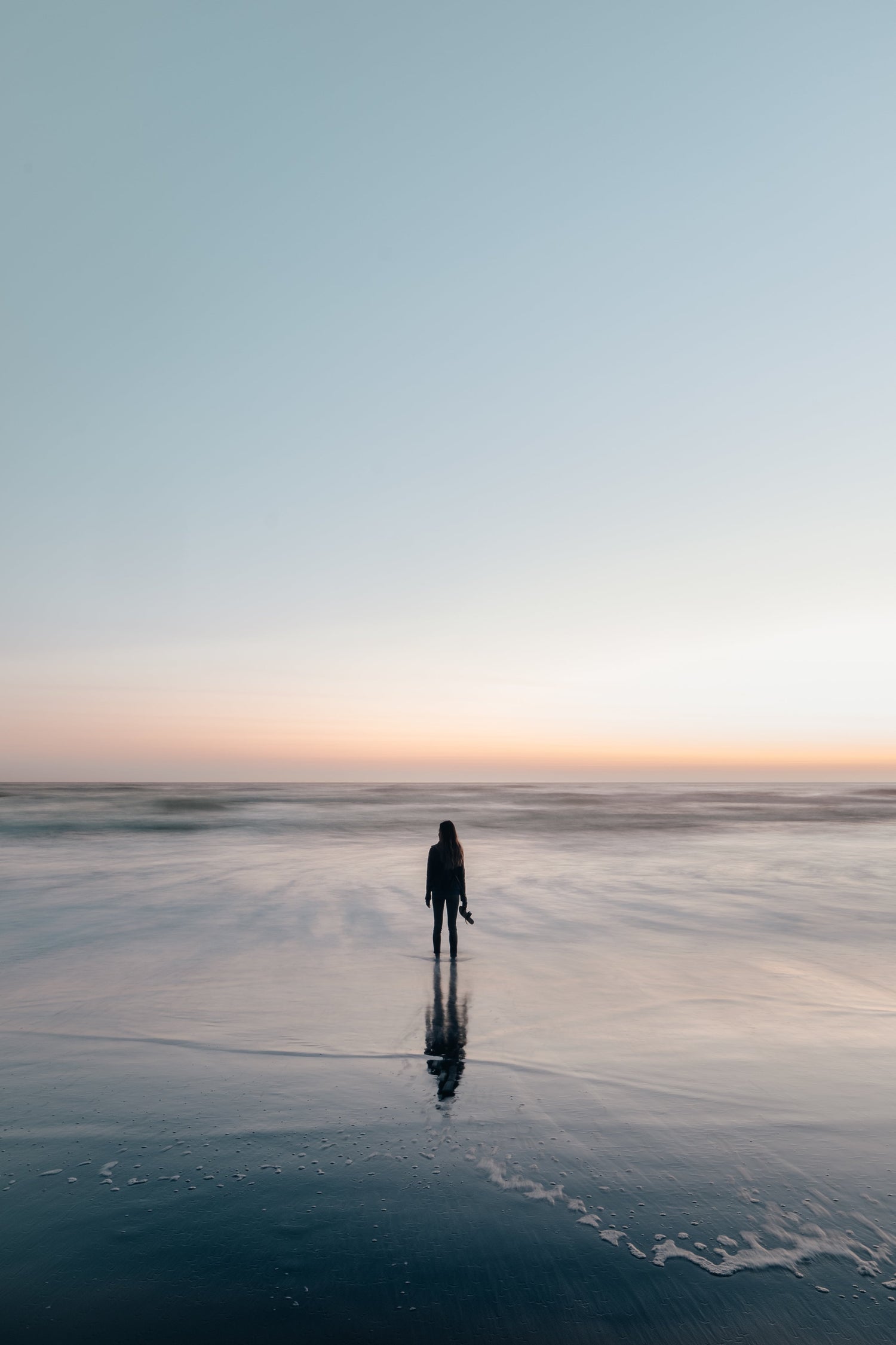 woman standing on the beach with waves covering her feet while she is holding her shoes in one hand while looking off into the distance