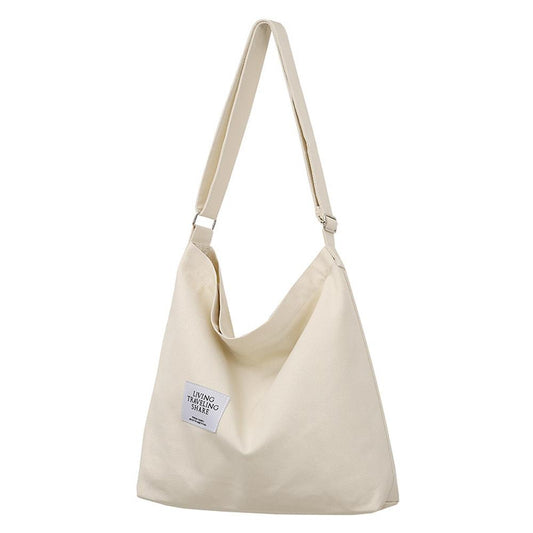 A minimalist cream-colored canvas crossbody handbag with an adjustable shoulder strap and a casual, relaxed design. The bag features a small label reading "Living, Traveling, Share" and is perfect for daily use.