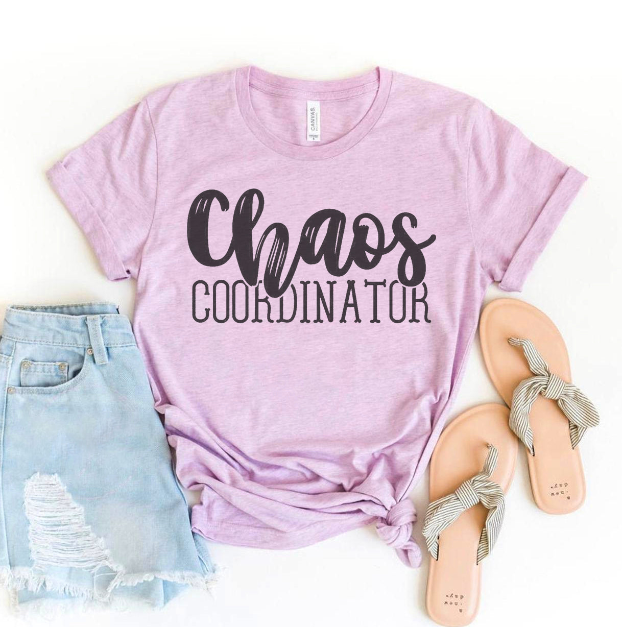 A light pink Bella Canvas t-shirt with the bold phrase "Chaos Coordinator" printed in black lettering, paired with light wash denim shorts and beige sandals. The shirt is styled in a laid-back, casual vibe, perfect for managing a busy life with style.