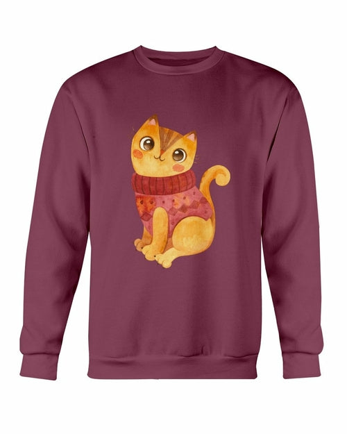 A maroon Gildan crewneck sweatshirt featuring an adorable illustration of a yellow-orange cat wearing a cozy red sweater. The sweatshirt showcases a soft, unisex design with a cute winter theme.