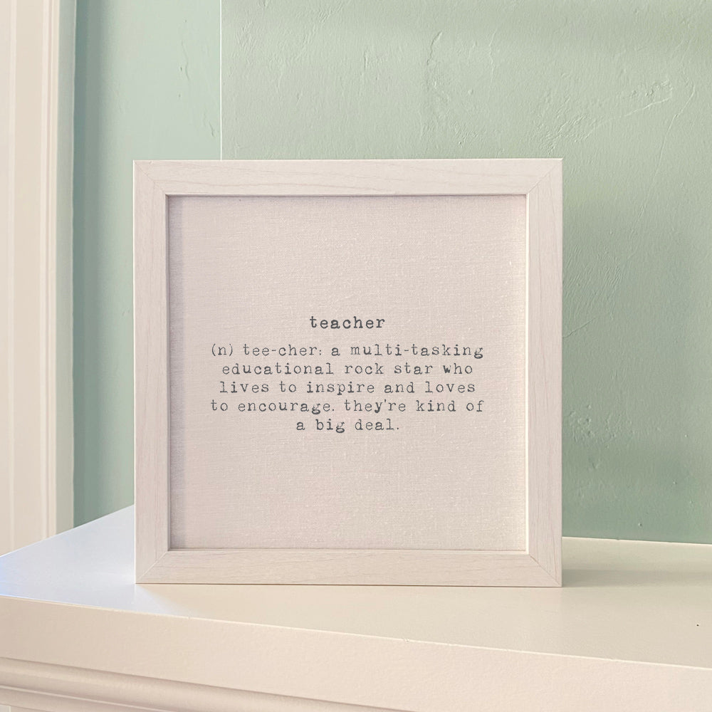 Framed decor print featuring the definition of a "teacher" in typewriter-style font, displayed on a natural, linen-style background with a whitewashed wooden frame. The text describes a teacher as an inspirational and encouraging figure.