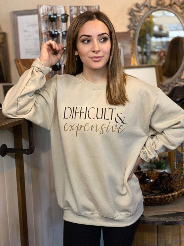 A young woman models a beige sweatshirt with the words "Difficult & Expensive" printed in bold black and gold script. The sweatshirt has a relaxed fit and is styled casually, perfect for a laid-back, confident look.