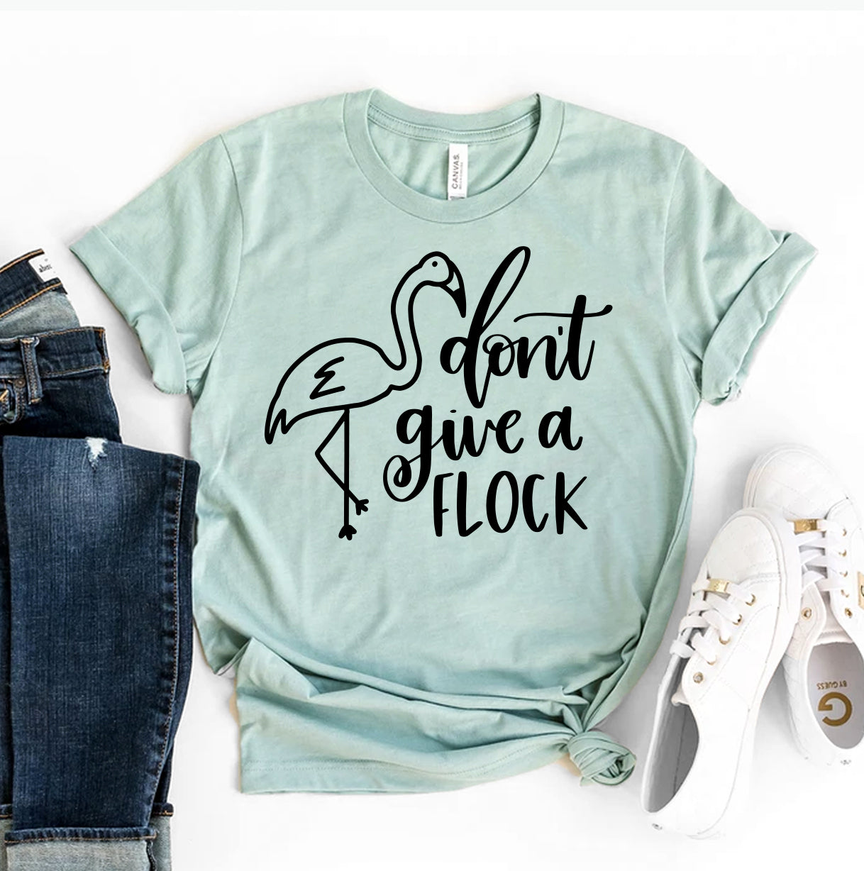 A mint green Bella Canvas t-shirt featuring a playful flamingo illustration with the words "Don't Give a Flock" in bold black script. The t-shirt is styled with rolled sleeves and paired with casual jeans and white sneakers.