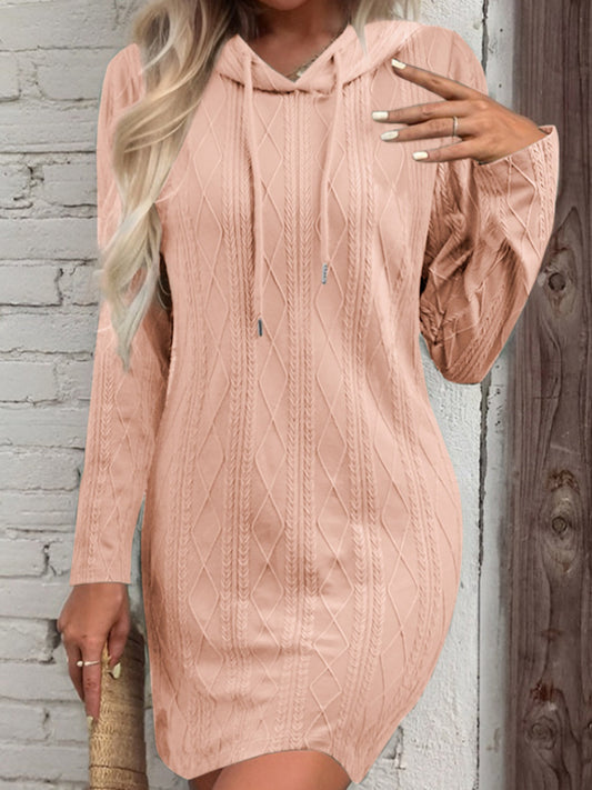 A stylish, light pink sweater dress featuring a hood and drawstring detail. The dress has a textured, cable-knit pattern and is paired with long sleeves. The cozy and casual look is perfect for cooler weather while maintaining a flattering and feminine silhouette.