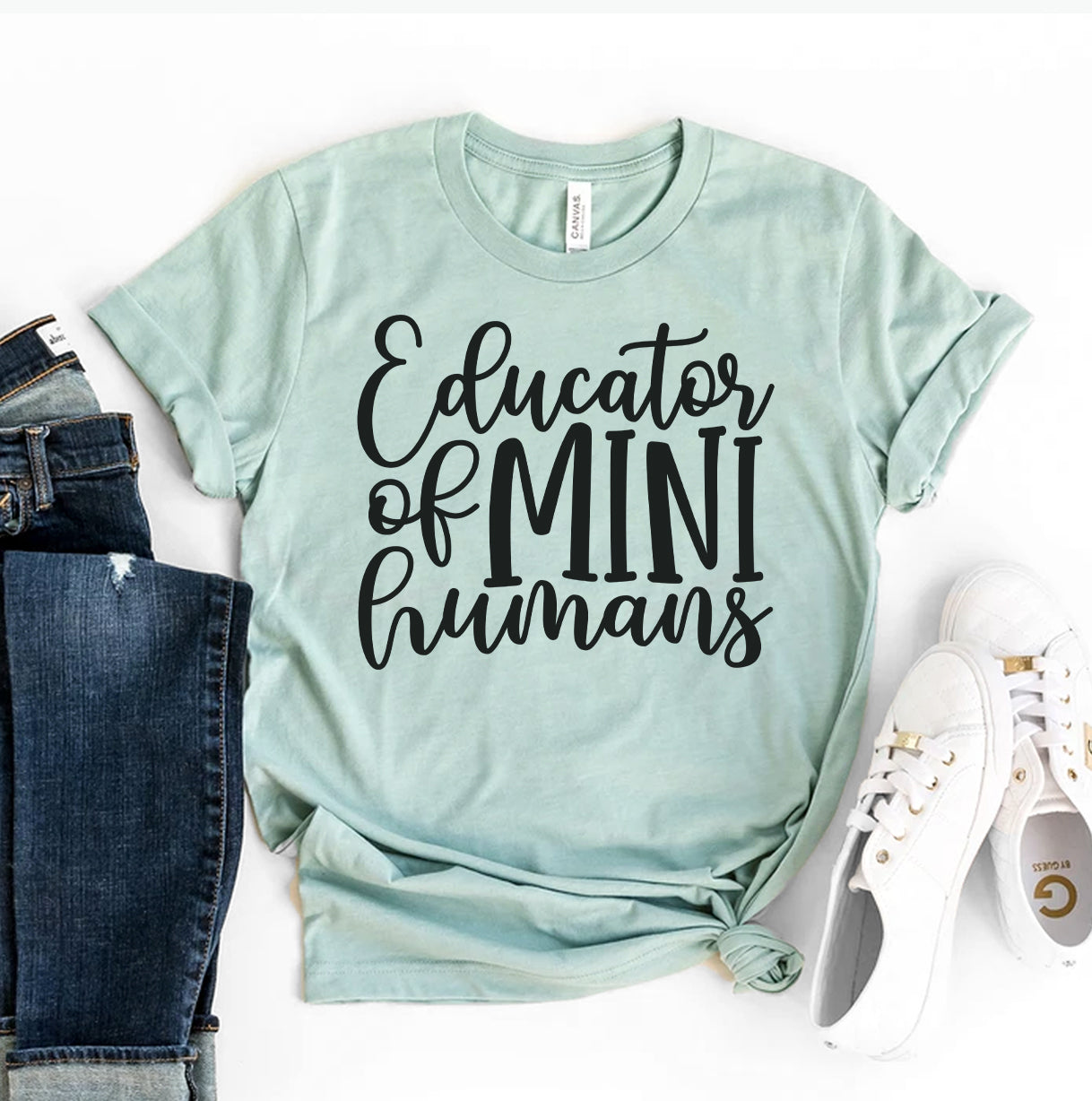 A light mint green Bella Canvas t-shirt with the bold black phrase "Educator of Mini Humans" printed across the chest. The shirt is styled with cuffed sleeves and paired with blue jeans and white sneakers, creating a casual and stylish look for teachers.