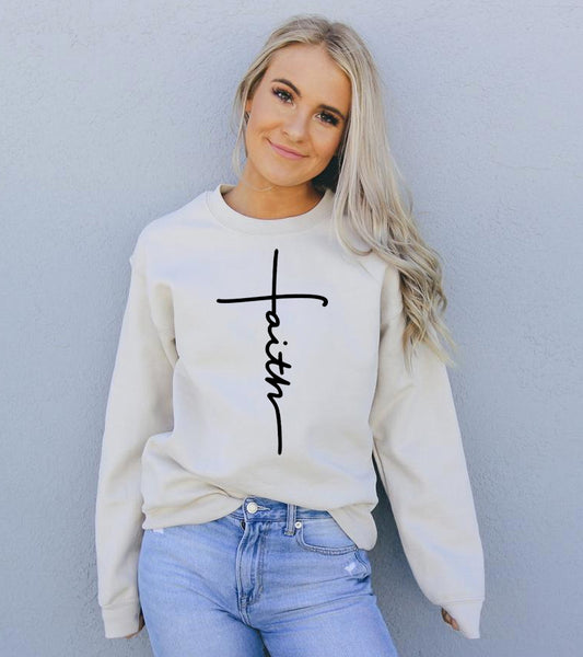 A woman with long blonde hair is wearing a light beige sweatshirt with a stylish black "Faith" graphic design in the shape of a cross. She pairs the sweatshirt with light-wash jeans and stands against a light gray wall, embodying a casual, comfortable, and faith-inspired look.