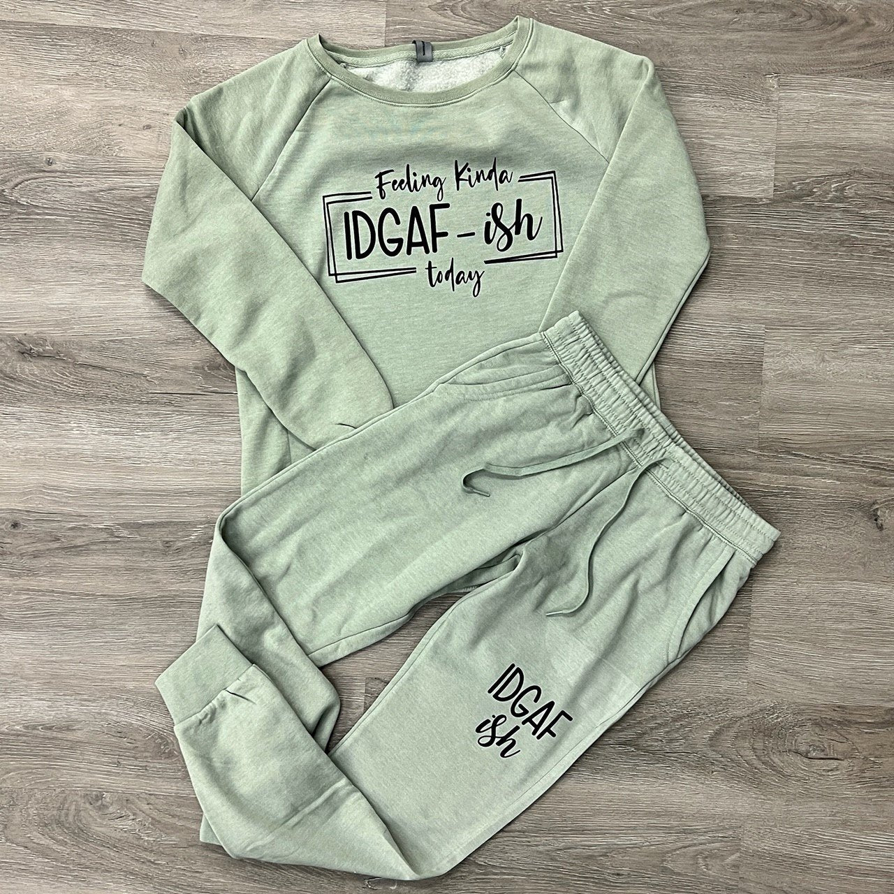 A light green sweat suit set is displayed on a wooden background. The top features a casual, long-sleeve design with the phrase "Feeling Kinda IDGAF-ish Today" printed across the chest. The matching joggers have "IDGAF-ish" printed on the left thigh. The cozy two-piece set is perfect for relaxed days with a fun, sassy vibe.