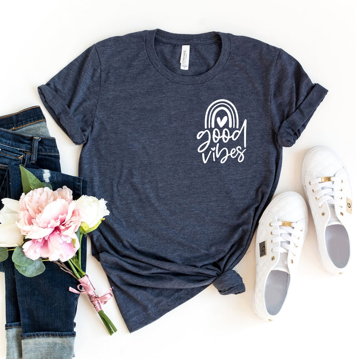 A dark gray Bella Canvas t-shirt featuring a minimalist white graphic that reads "Good Vibes" with a simple rainbow and heart design. The shirt is styled with rolled sleeves, paired with distressed denim jeans, white sneakers, and a bouquet of peonies, giving off a casual and cheerful vibe.