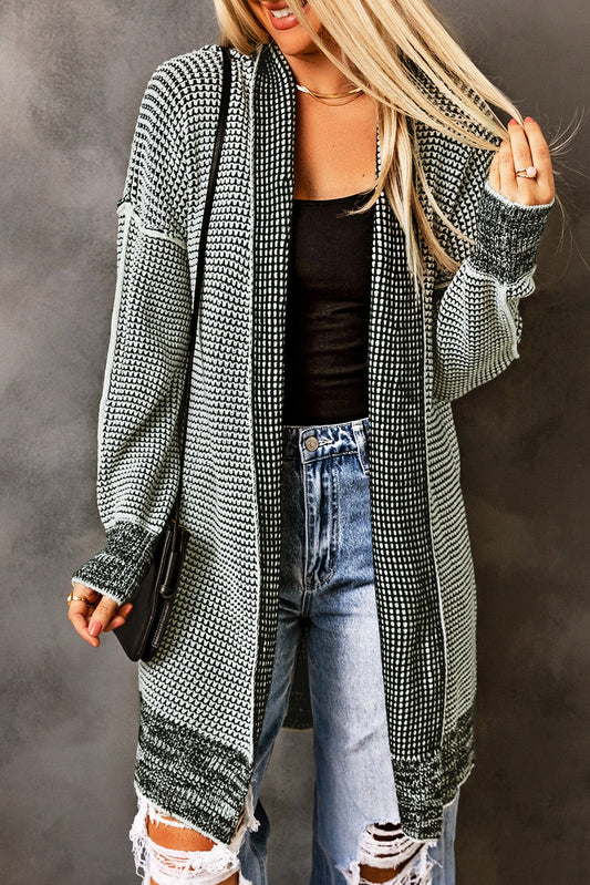 A woman wearing a cozy, oversized gray plaid knitted long open front cardigan. The cardigan features a plaid knit pattern with a relaxed fit and is styled with a black tank top and distressed light-wash jeans.