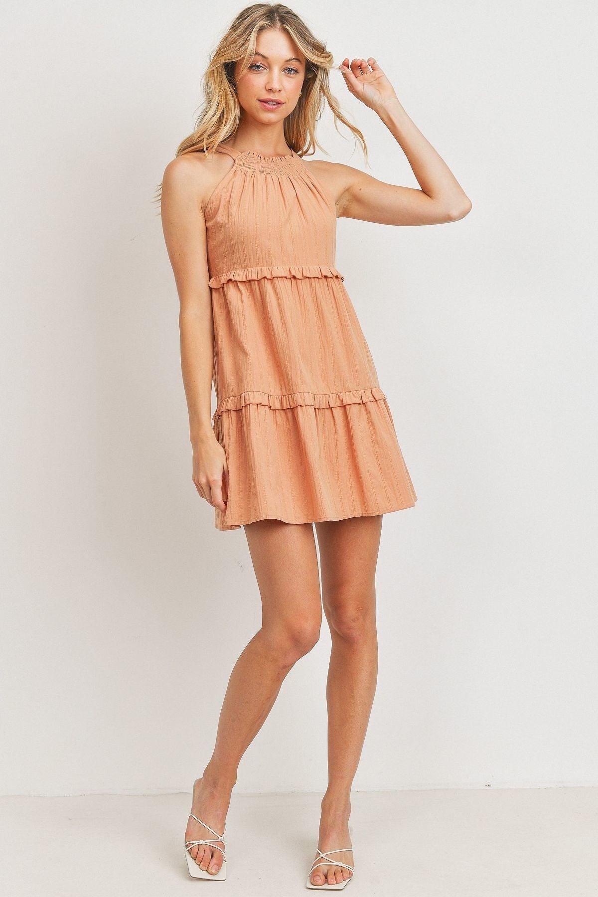 A woman wearing a Halter Top Tiered Dress in a soft peach tone. The dress features a high halter neckline, tiered ruffle layers, and a flowy silhouette, perfect for a breezy summer look.