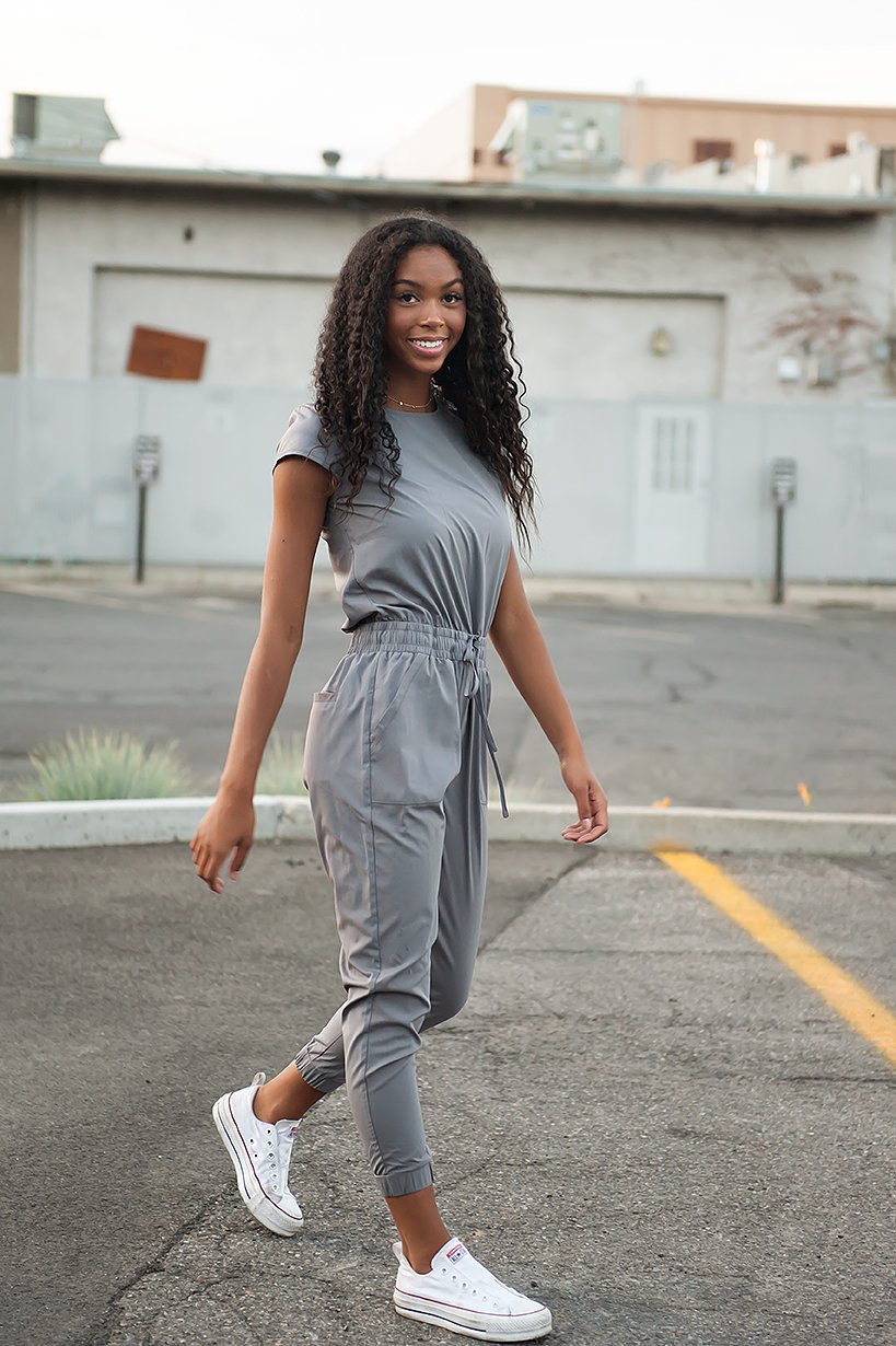 A young woman is wearing a Cement-colored Jumpsuit with a relaxed fit. The jumpsuit features short sleeves, a cinched waist with a drawstring, and cuffed jogger-style pants. She completes her casual look with white sneakers, creating an effortlessly stylish and comfortable outfit perfect for day-to-day wear.