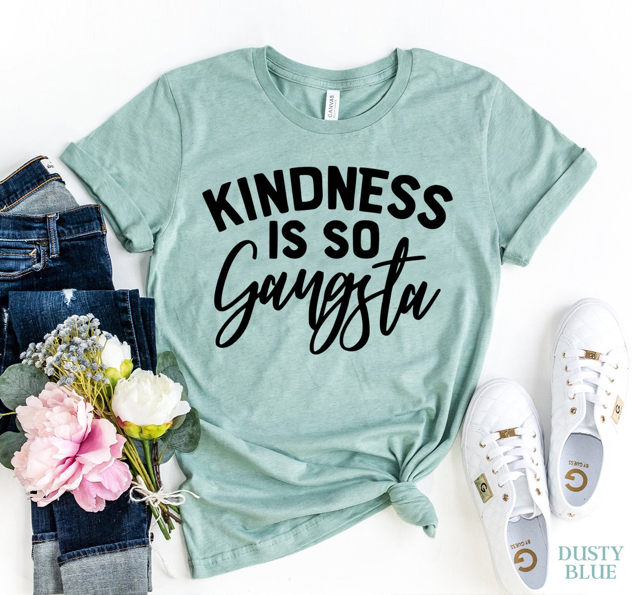 A flat lay of a light blue "Kindness Is So Gangsta" T-shirt styled with dark blue jeans, white sneakers, and a bouquet of flowers. The shirt has a bold black statement print on the front and is tied at the bottom for a casual chic look. The brand is Bella Canvas.