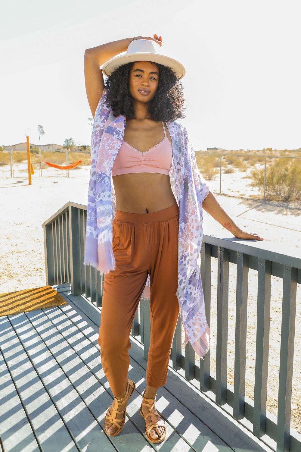 A woman poses outdoors in a light, flowing kimono with a colorful splatter design. The kimono is a blend of soft lavender, pink, and white hues with a relaxed fit. She pairs it with a peachy bralette, rust-colored flowy pants, and sandals, accessorized with a wide-brimmed hat.